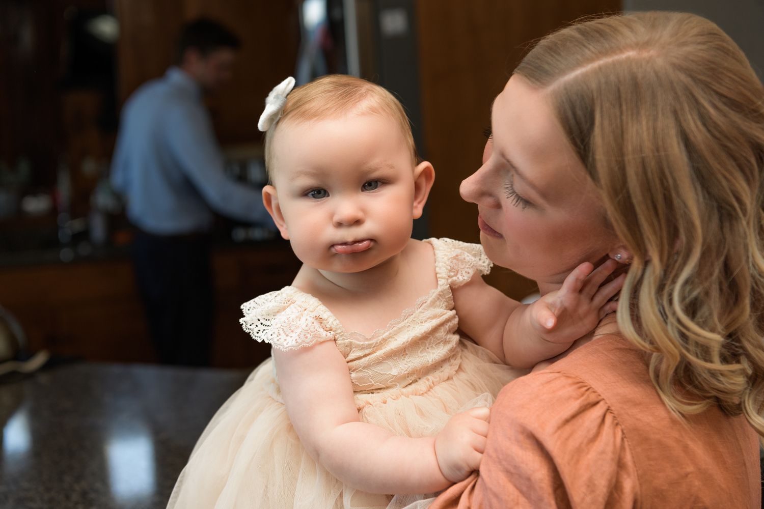Baby in cream-colored dress being held during wedding celebration.