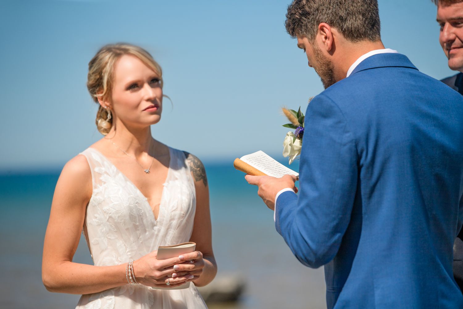A wedding ceremony takes place on a beach with a bride in white and officiant in blue exchanging vows.
