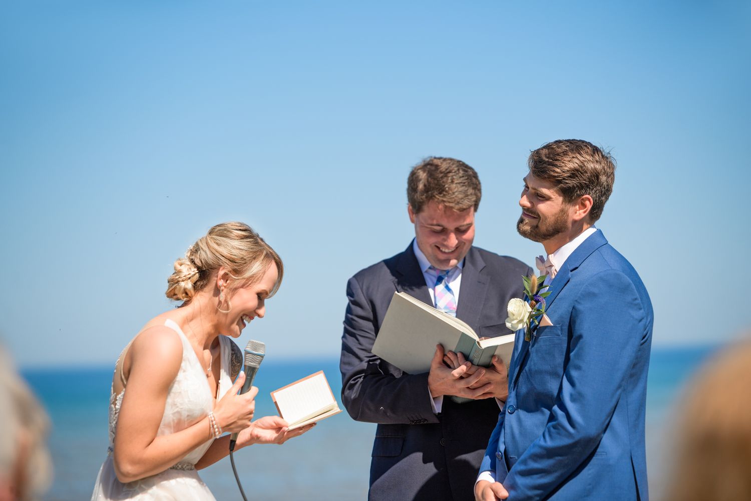 A seaside wedding ceremony with participants reading from books against a bright blue sky.
