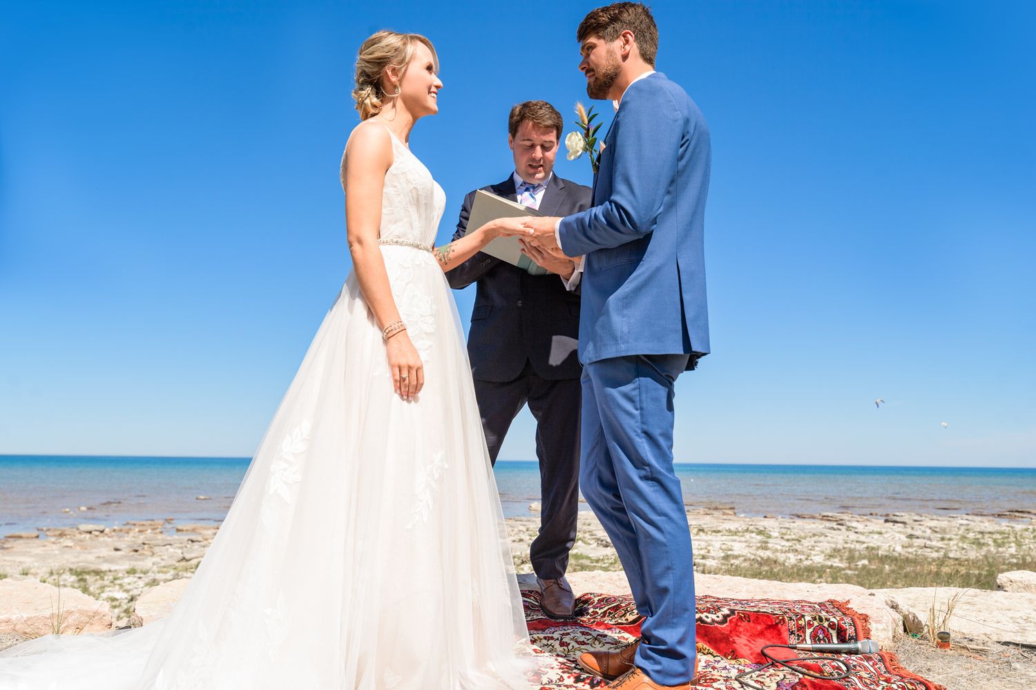 An intimate beach wedding ceremony with participants gathered on a decorative red rug.