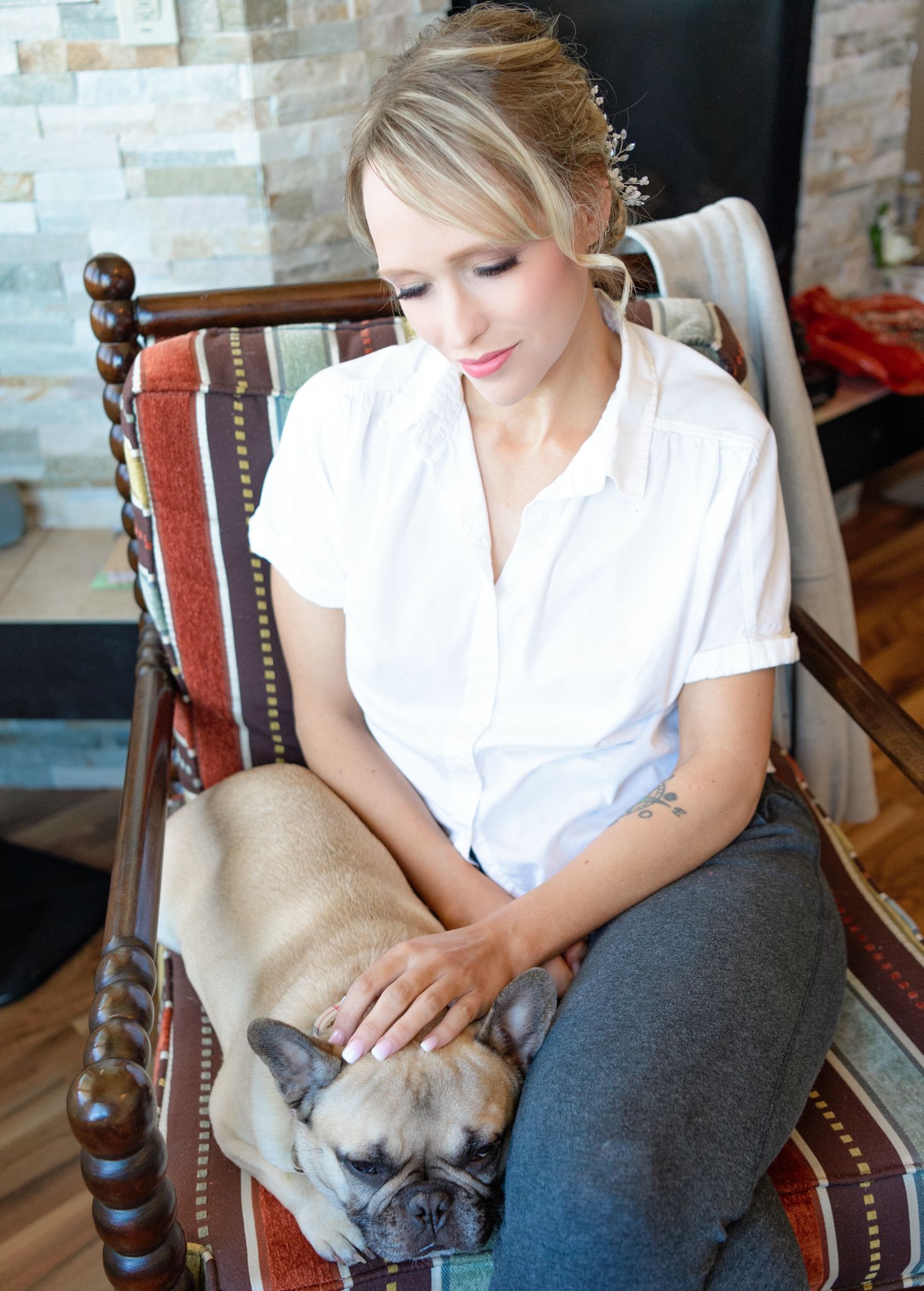 Woman in white shirt sits in vintage chair while petting sleeping French Bulldog.
