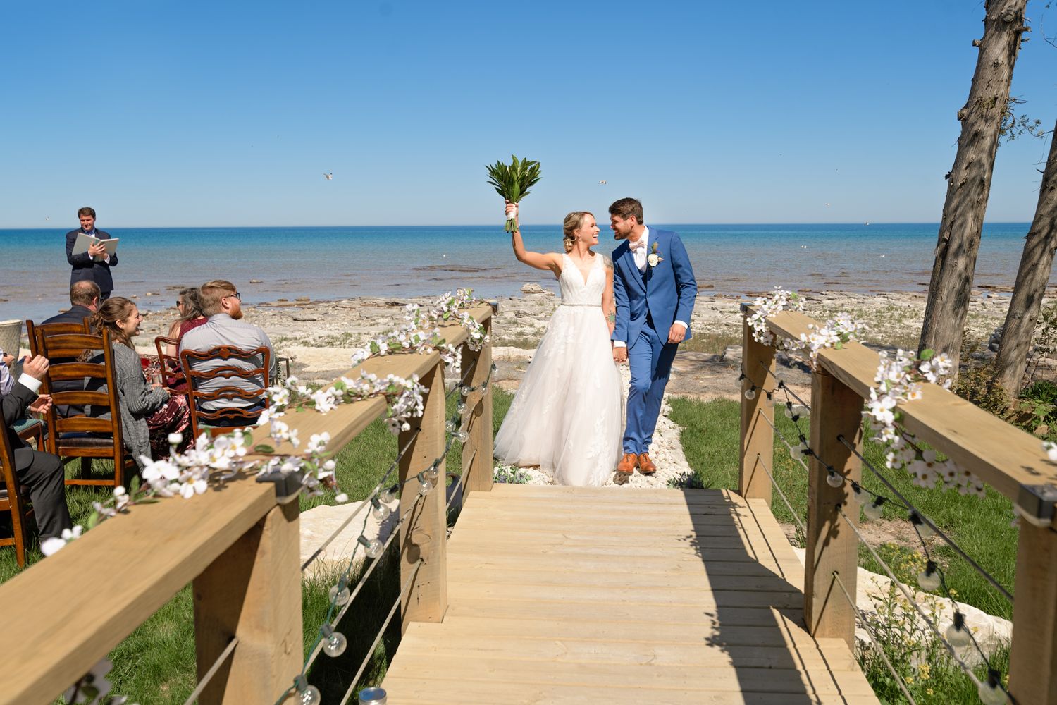 Wedding couple walks down wooden boardwalk decorated with greenery overlooking ocean.