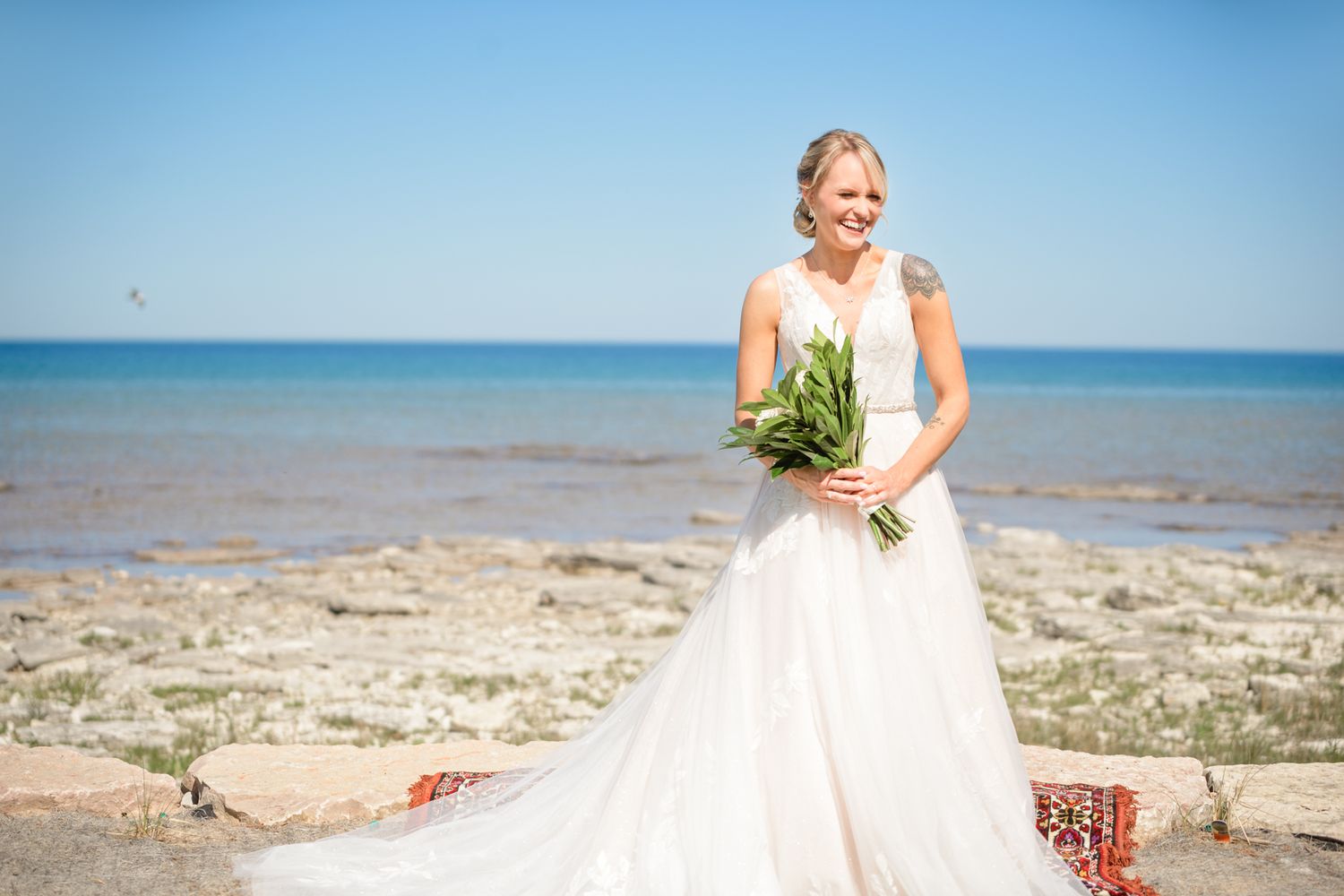 A bride in a flowing white dress stands on a rocky beach holding a green and white bouquet against a blue ocean backdrop.