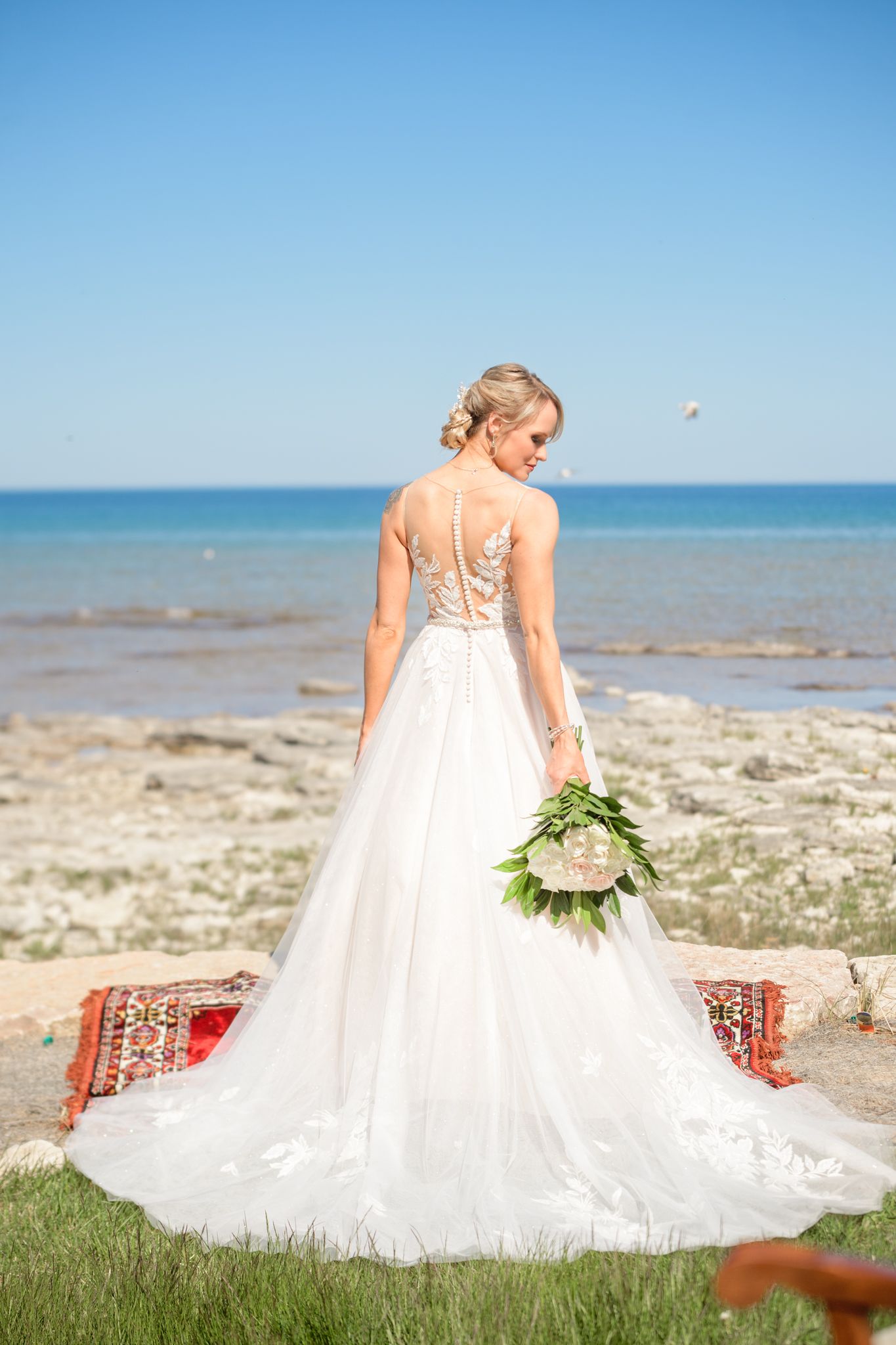 Elegant bridal gown with sheer back detail and flowing train photographed against ocean backdrop.