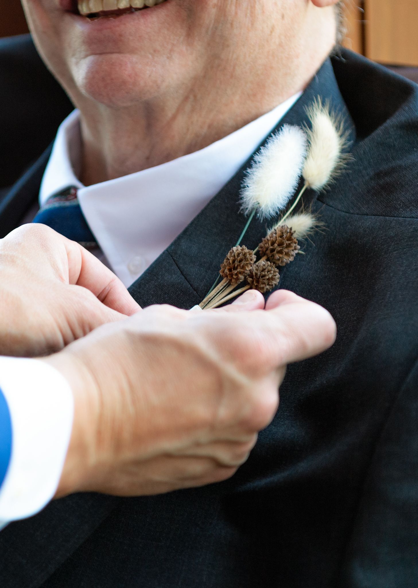 Close up of hands pinning a rustic boutonniere with pinecones and white feathers to a black suit lapel.