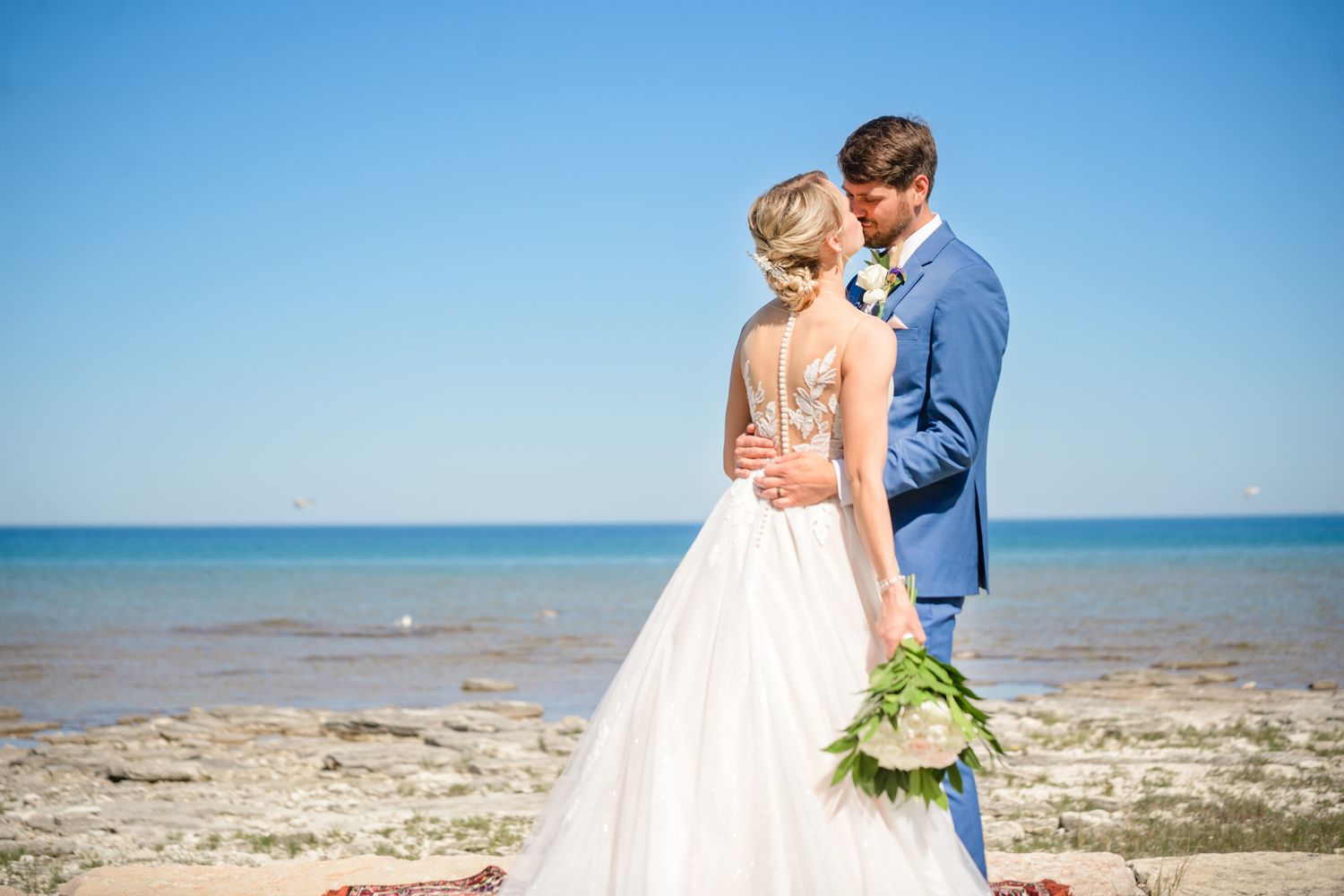 Waterfront wedding portrait with couple in blue suit and white dress against ocean backdrop.