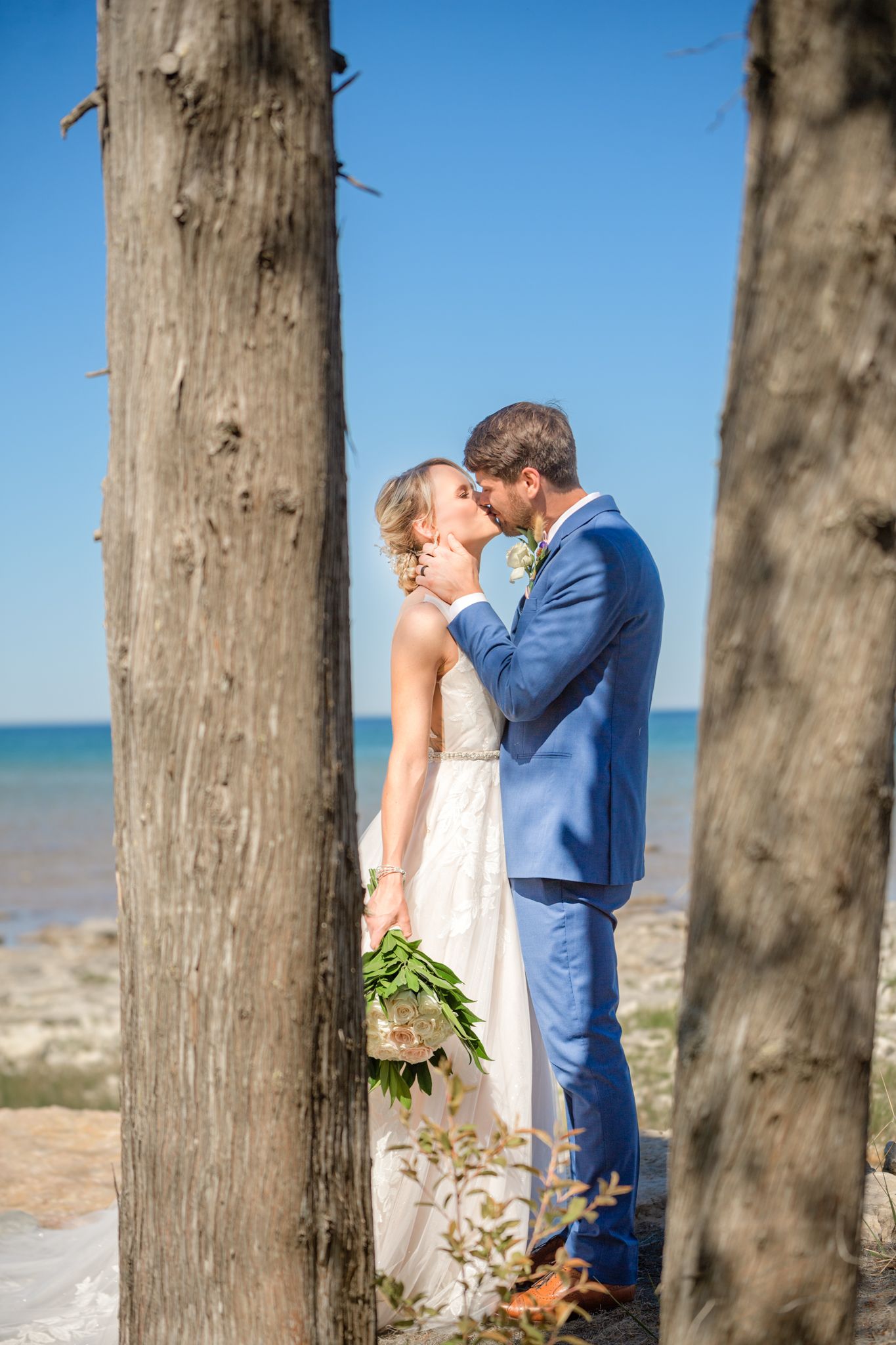 Wedding couple shares intimate moment between two tall trees with ocean in background.