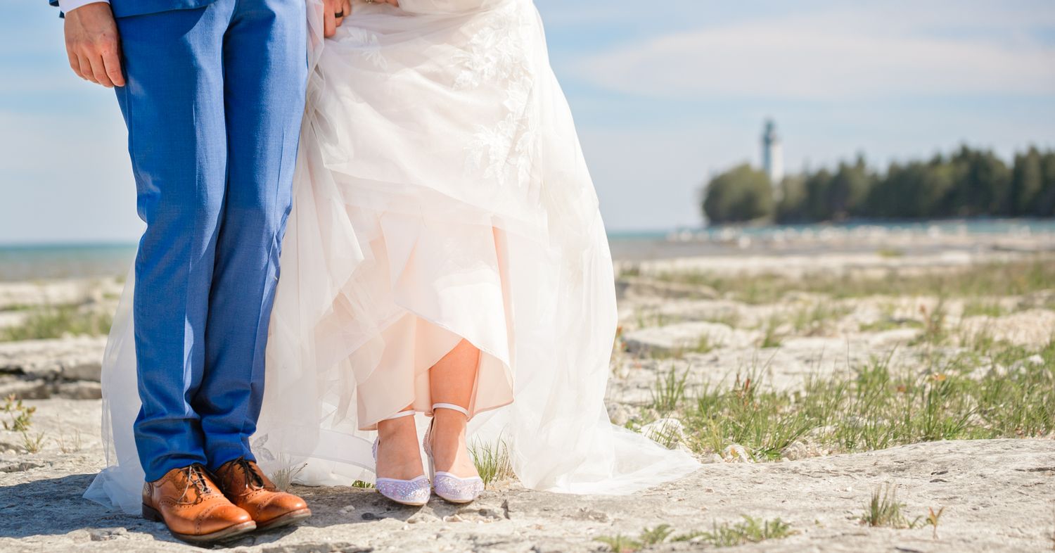 Detail shot of bride and groom's feet on sandy beach with wedding dress hem visible.
