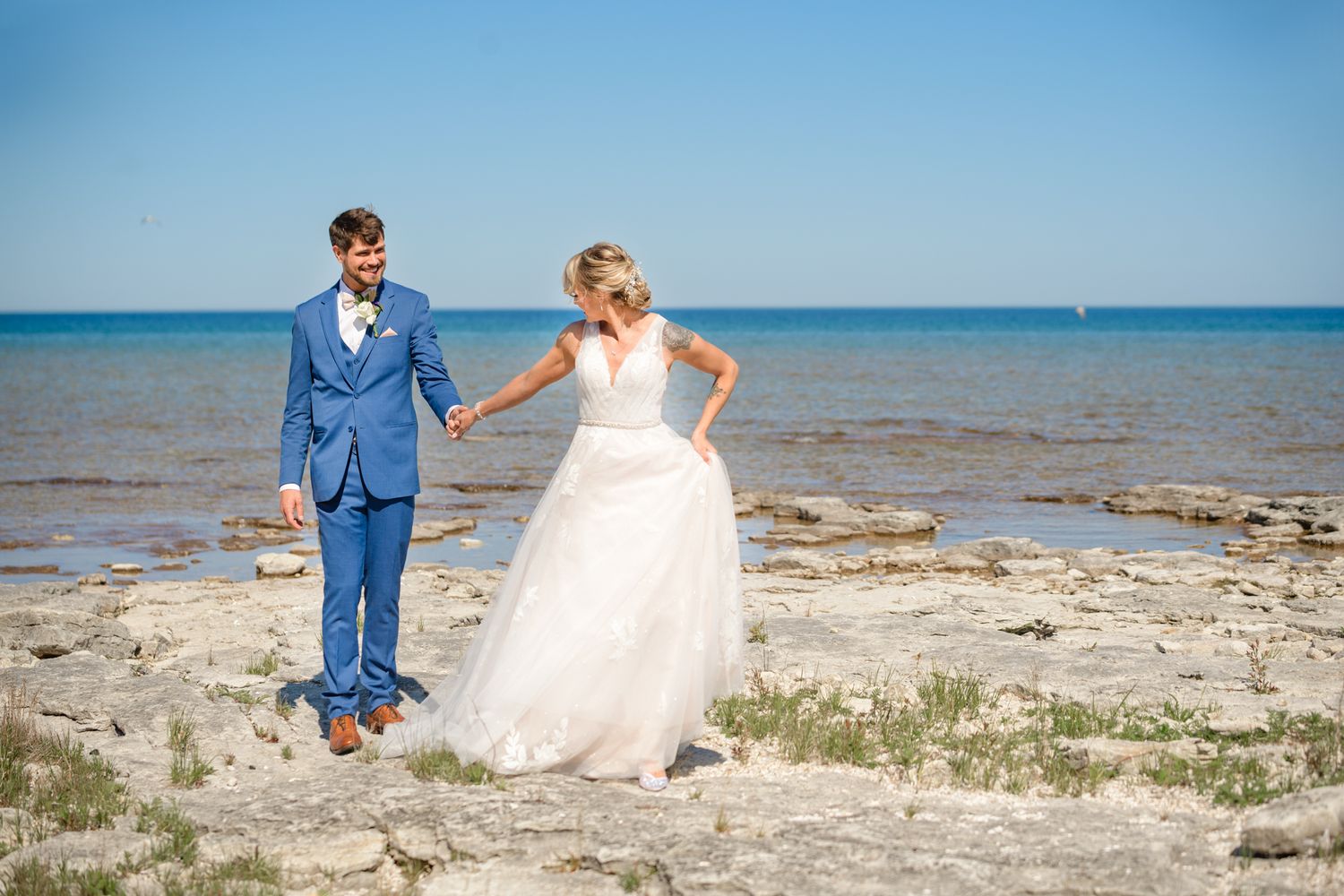 A wedding couple stands together on a scenic beach with rocky outcrops and clear blue water.
