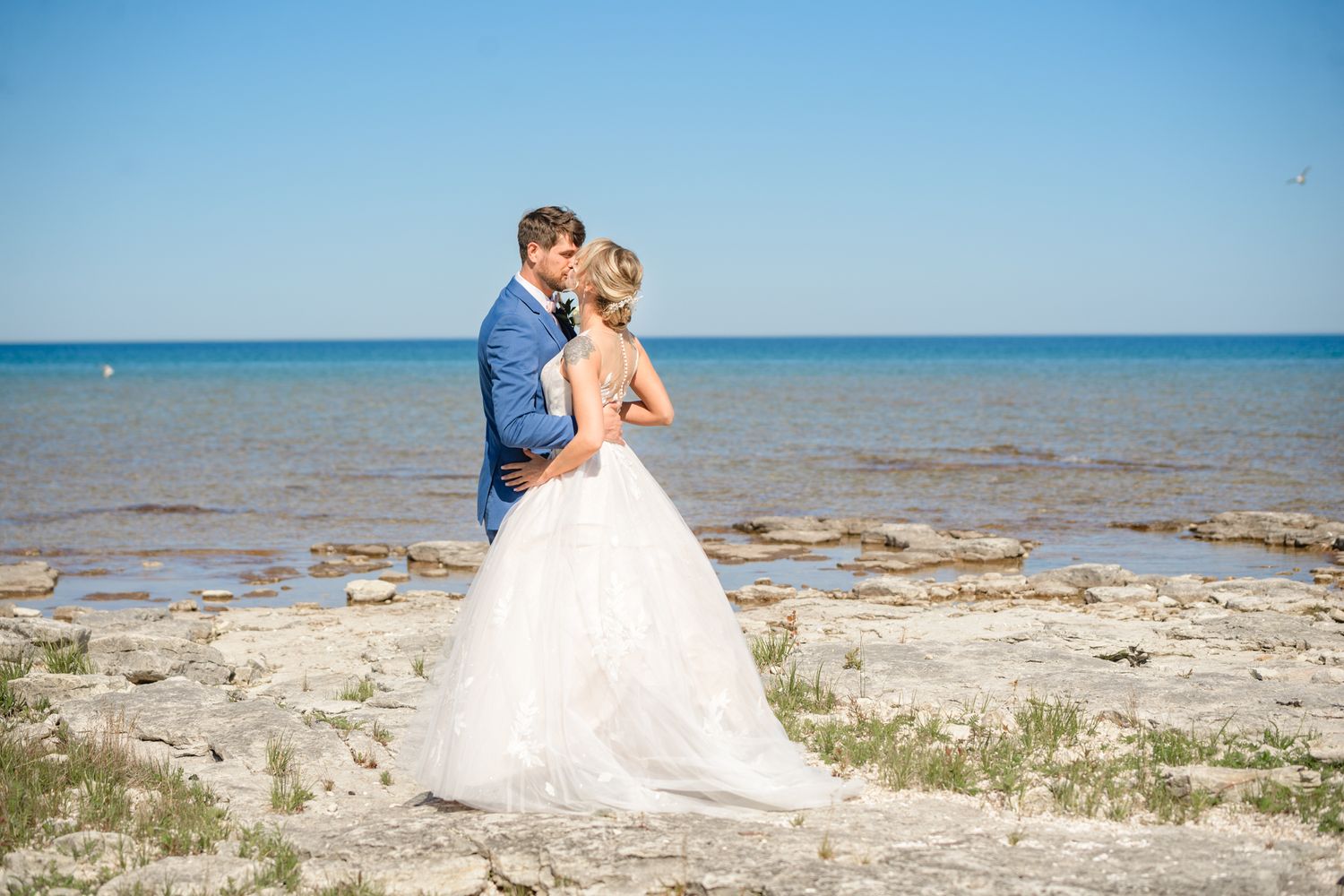 A romantic beach wedding scene with a couple embracing on a rocky shoreline against a bright blue ocean backdrop.