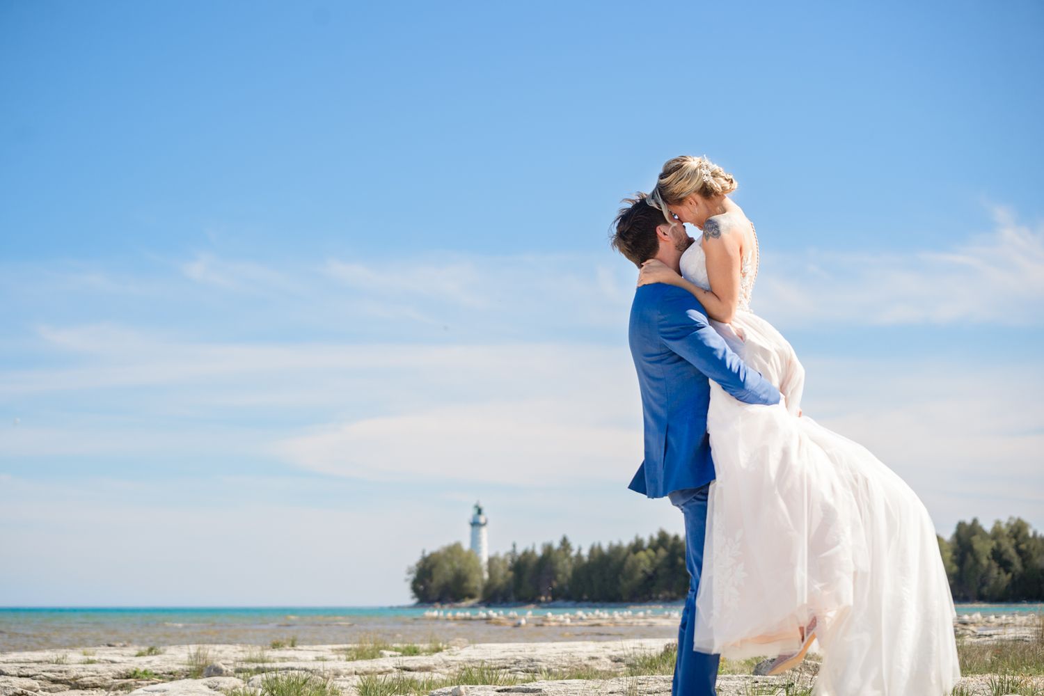 A joyful wedding lift pose on a beach with a lighthouse and trees in the distance.