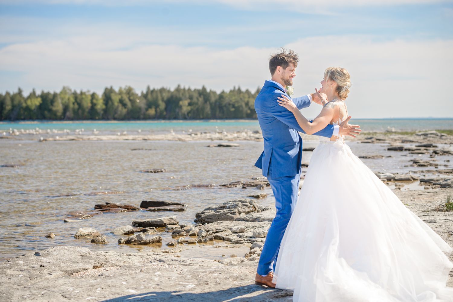 A wedding couple dances on a rocky beach shoreline with forest and ocean in the background.