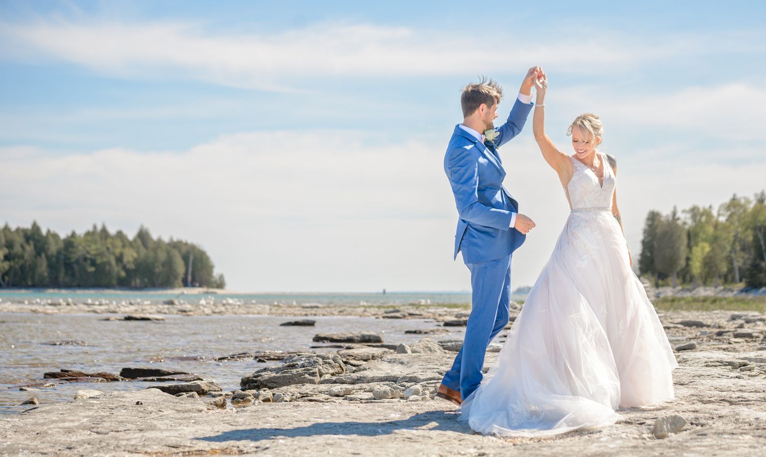 A couple in wedding attire dancing on a rocky beach with forest and water in the background.