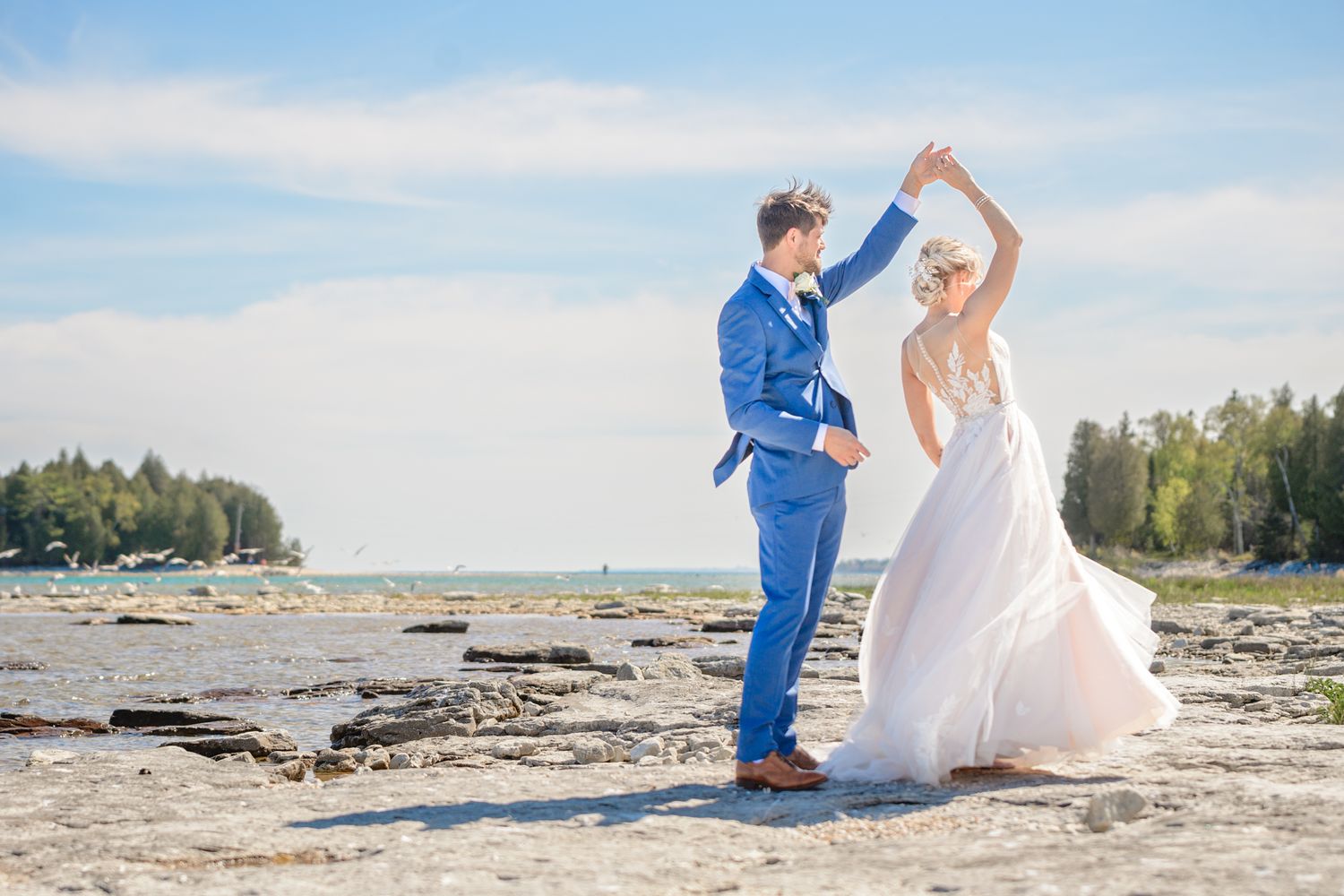 Wedding couple dancing elegantly on a beach shoreline with evergreen trees in the background.