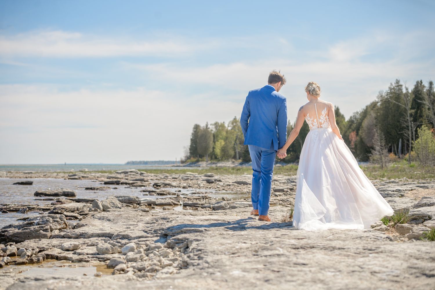 A couple in wedding attire walking along a rocky coastal shoreline.