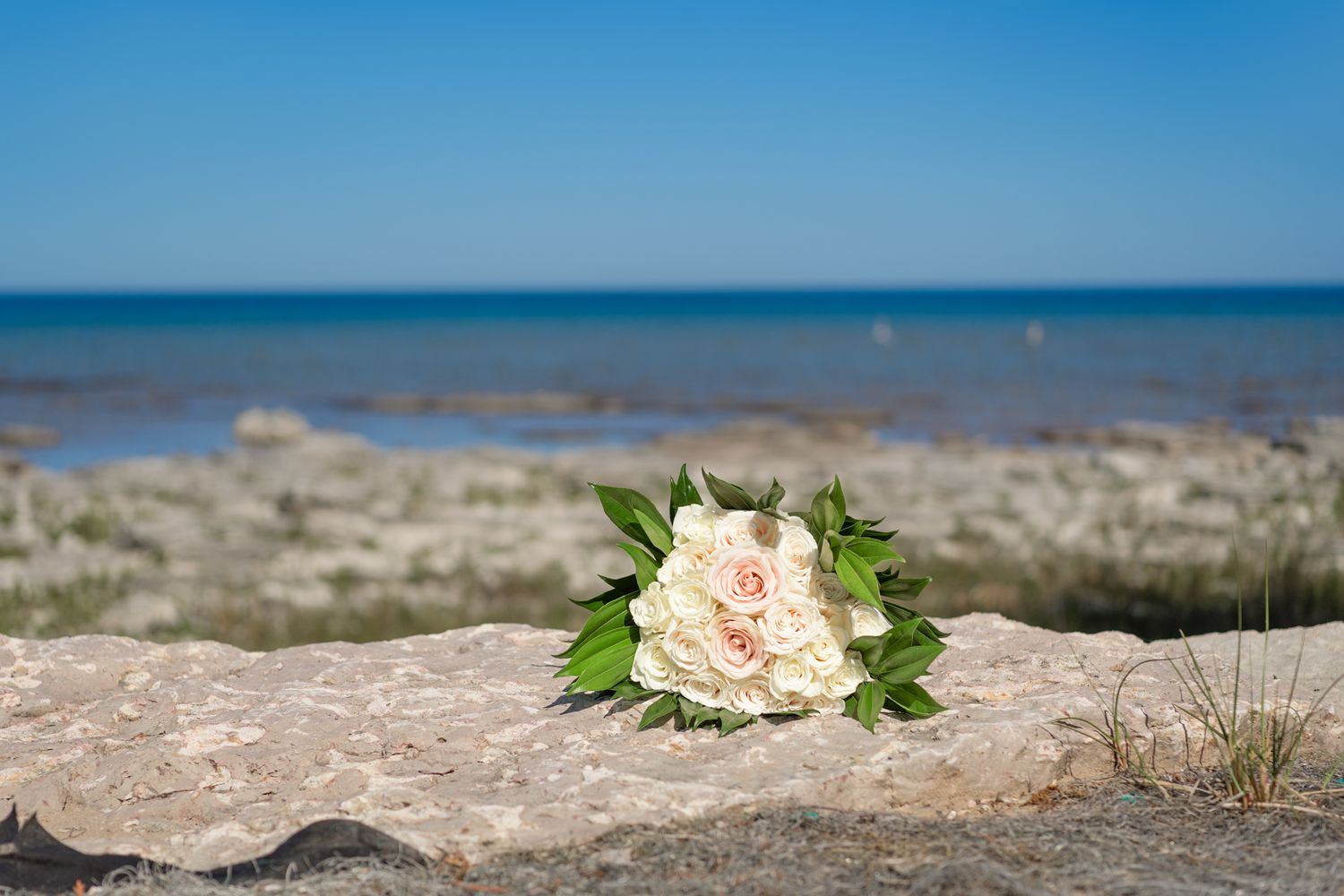 A delicate pink and white bridal bouquet rests on a rocky beach shore with blue ocean waters in the background.