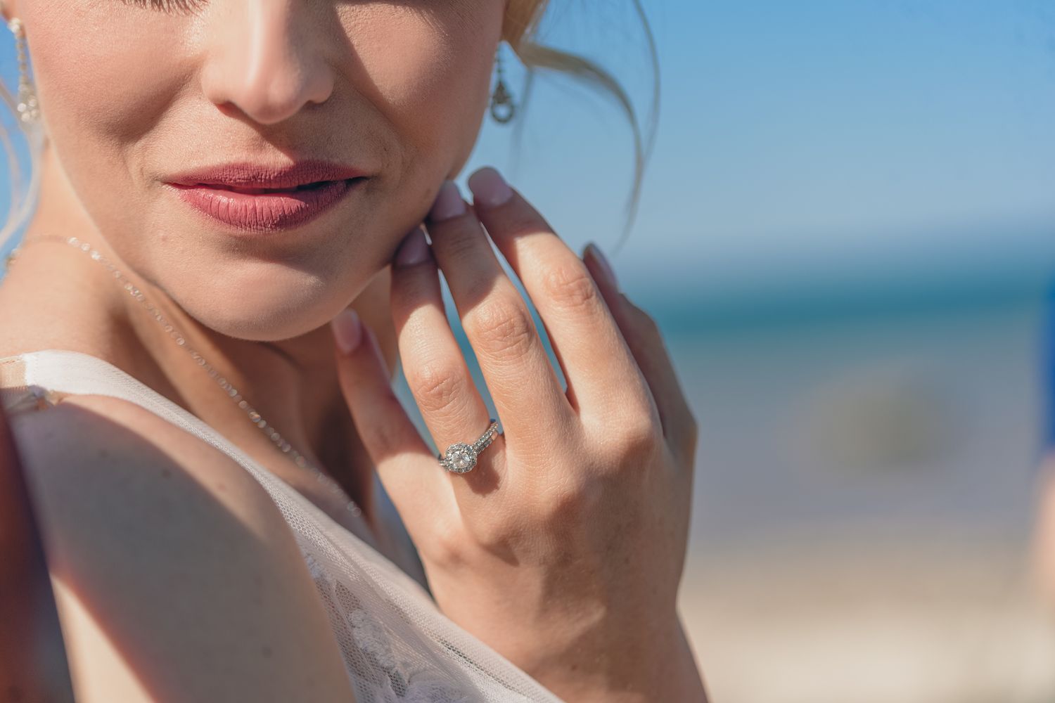 Close-up detail of a diamond engagement ring and manicured hand against a beach backdrop.