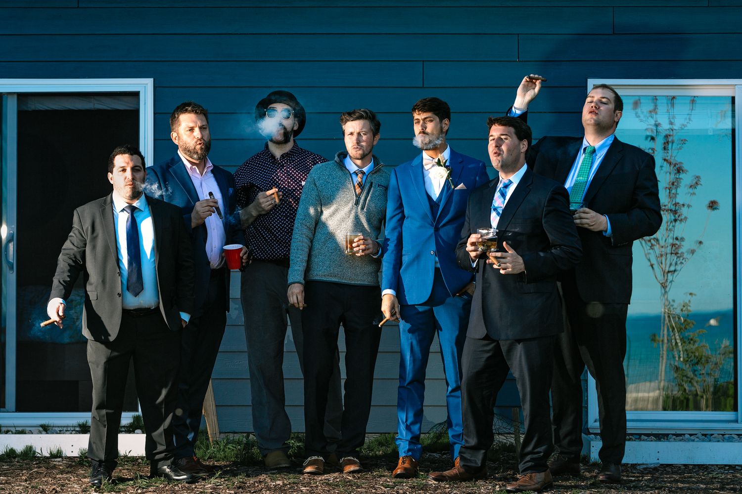 A group of groomsmen in formal attire pose together at dusk with blue lighting.