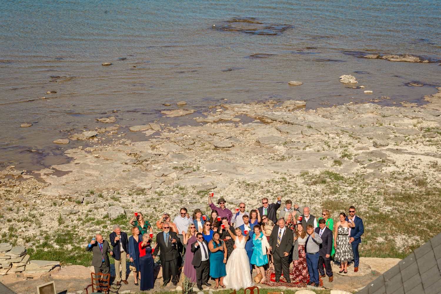 Aerial view of wedding guests assembled for a group photo on a rocky beach overlook.