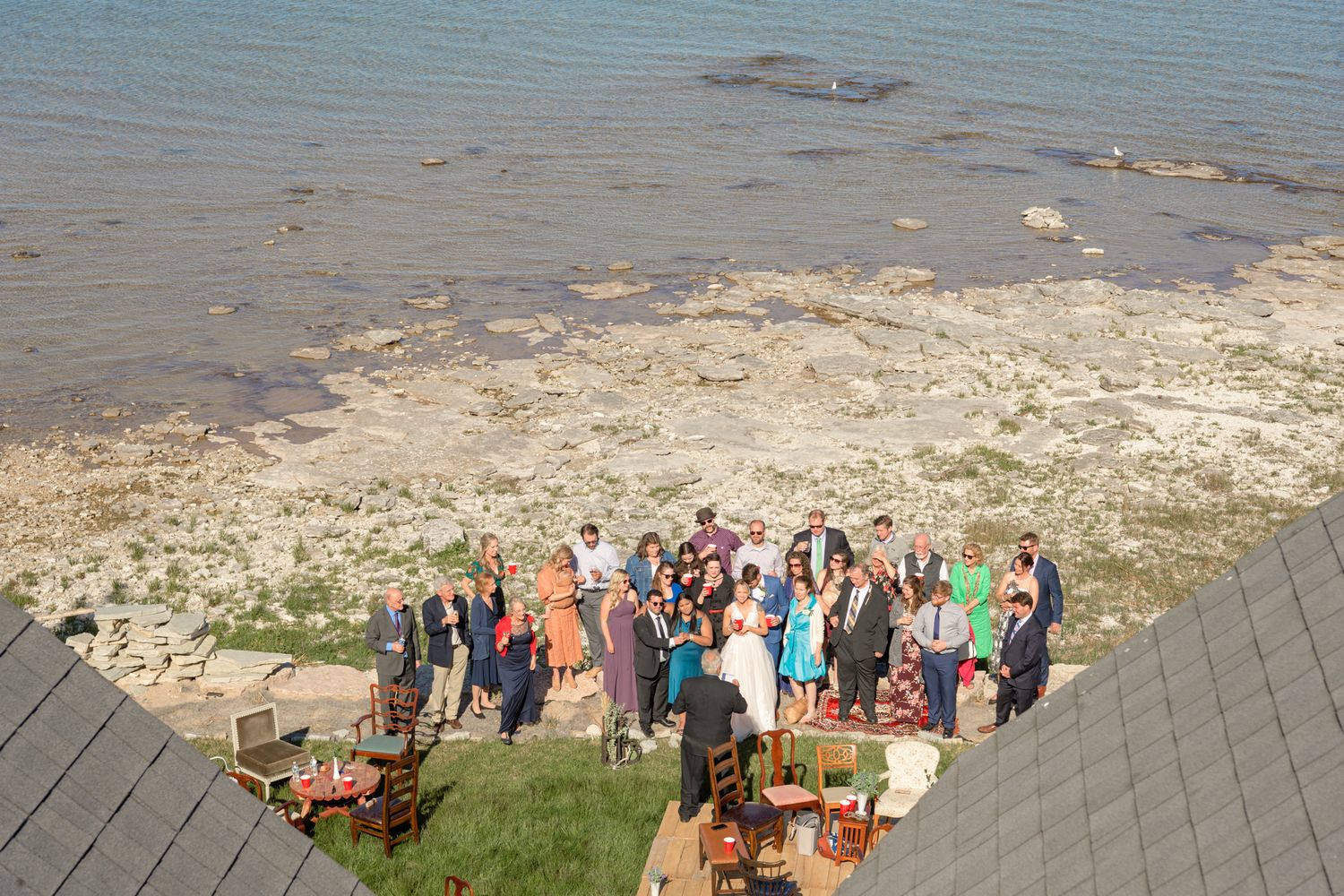 Bird's eye view of wedding celebration with guests gathered on coastal cliff near outdoor furniture.