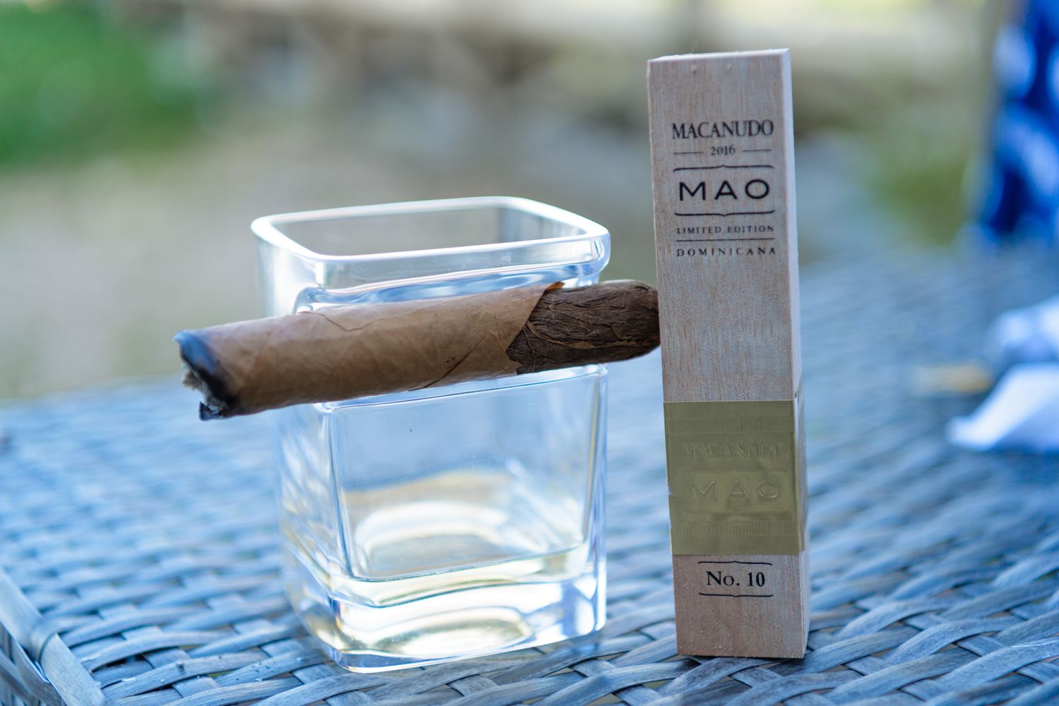 Glass tumbler with cigar and gray product package on blue table outdoors.