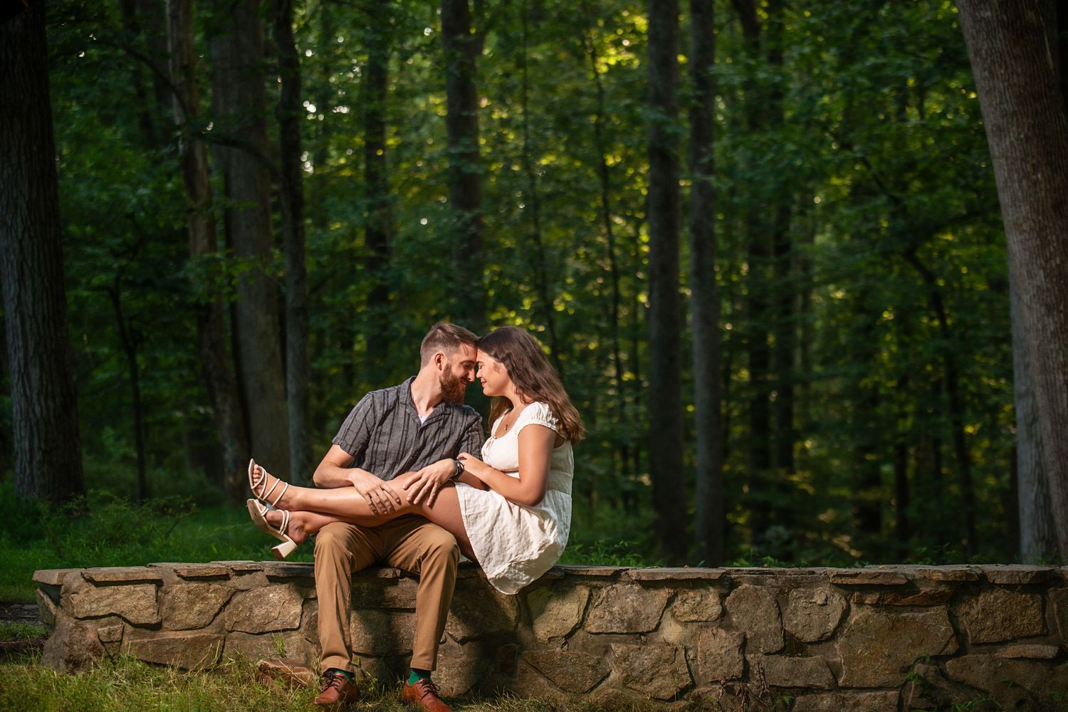 A romantic couple sits together on a rustic stone wall surrounded by lush forest greenery at sunset.