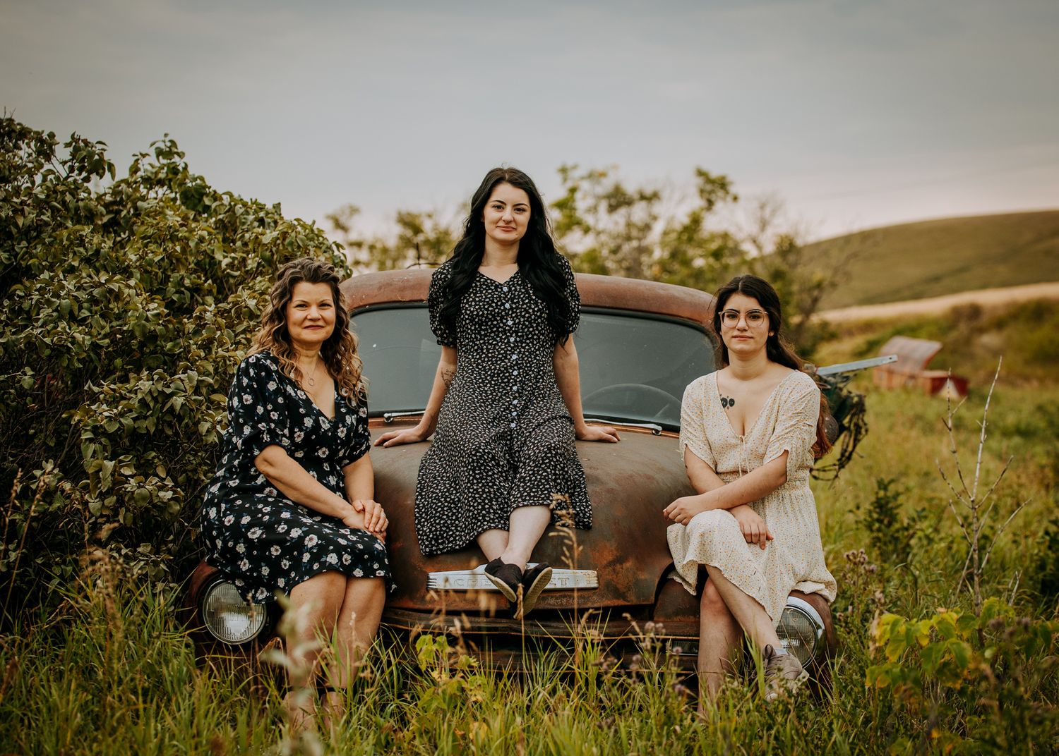 Three women in vintage dresses pose on classic car in rural setting.