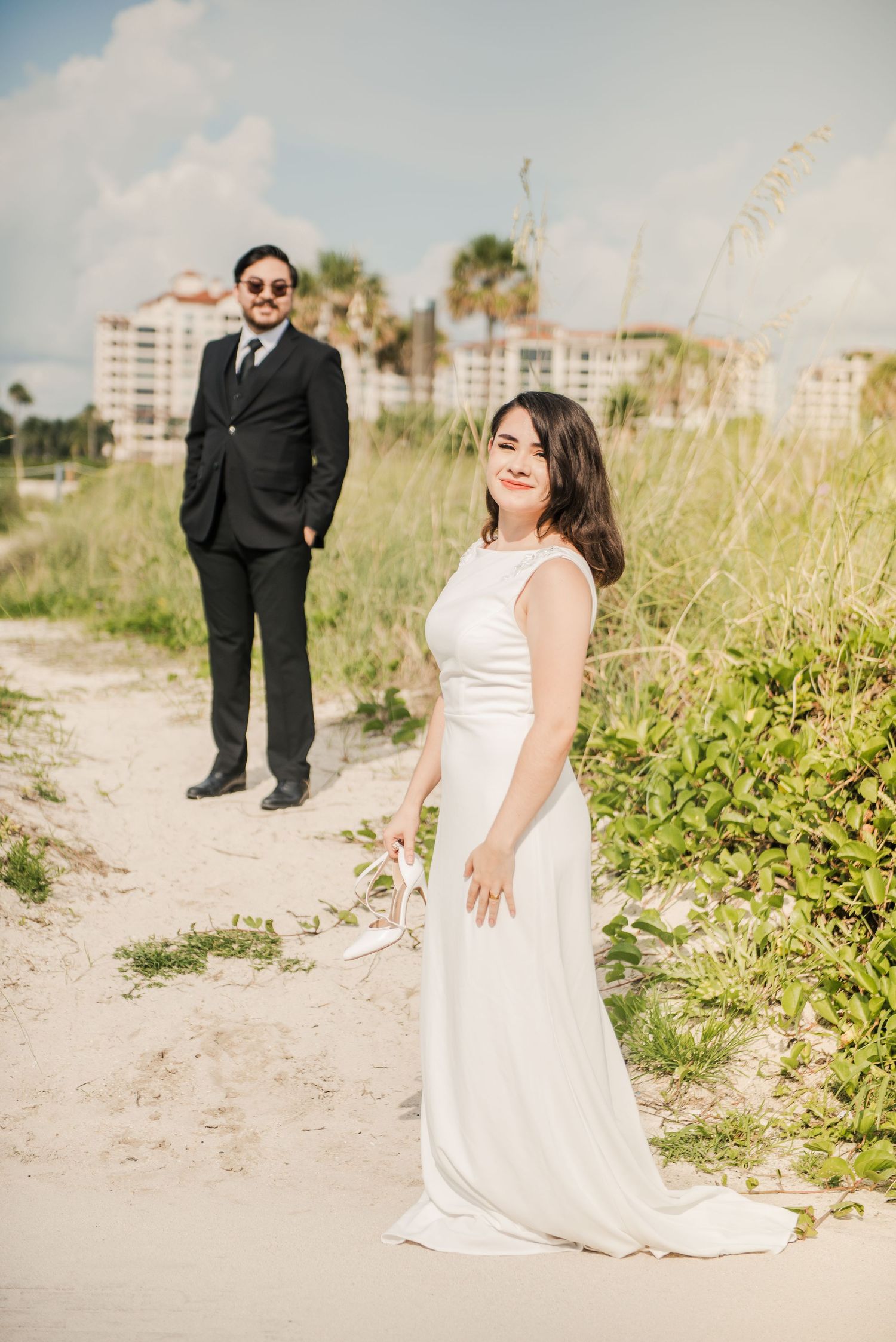 Bride in flowing white dress and groom in black suit pose on sandy beach path with coastal buildings in background.