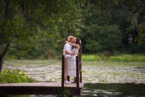 A couple embraces on a wooden dock overlooking a serene pond surrounded by lush greenery.