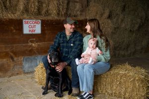 A family sits on hay bales in a rustic barn setting with their black dog and baby, near a 'Second Cut Hay' sign.