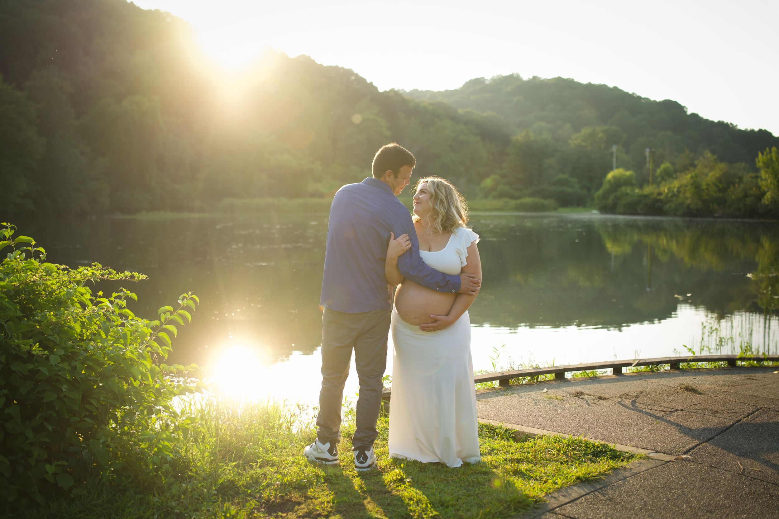 A couple stands silhouetted against a golden sunset by a serene lake as sunlight filters through trees.