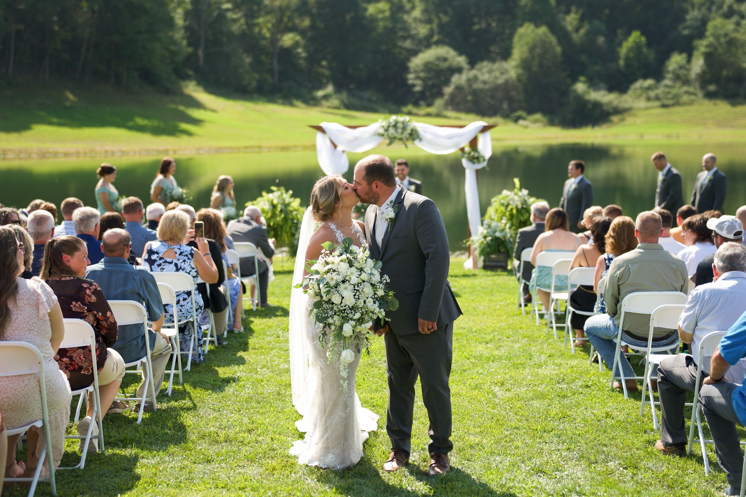 A beautiful outdoor wedding ceremony takes place on a sunlit lawn surrounded by guests in white chairs with forest scenery.
