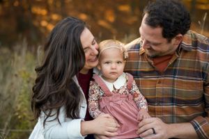 A family shares a tender moment together outdoors during autumn, with their baby wearing a pink jumper dress.