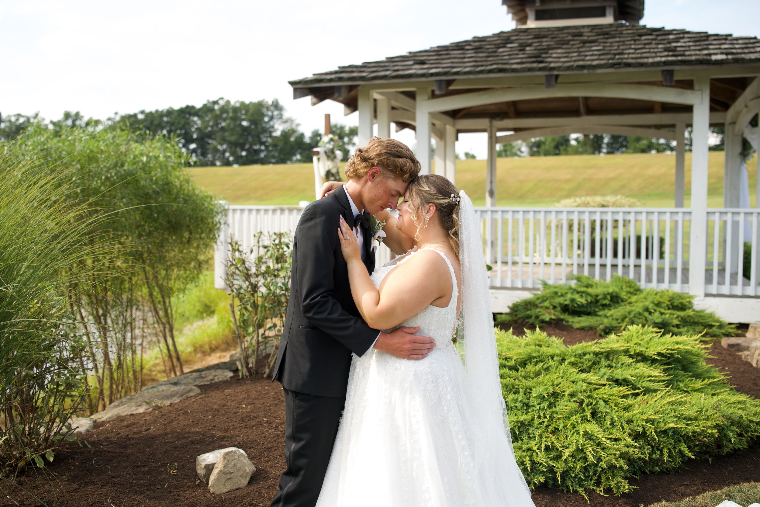 A romantic couple shares a tender moment in a white gazebo surrounded by lush greenery and gardens.