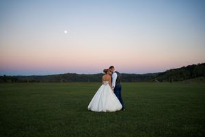 A couple shares a romantic moment in an open green field during sunset with a soft pink sky in the background.