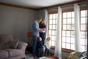 A couple stands together by large French doors in a cozy living room with white curtains and grey furniture.