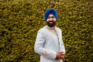 Sikh groom in blue turban and white sherwani standing against green hedge backdrop.