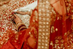 Close-up of traditional Indian bridal hands with intricate henna designs against red embroidered fabric.