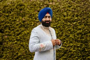 Groom in traditional Sikh attire smiling against green hedge background.
