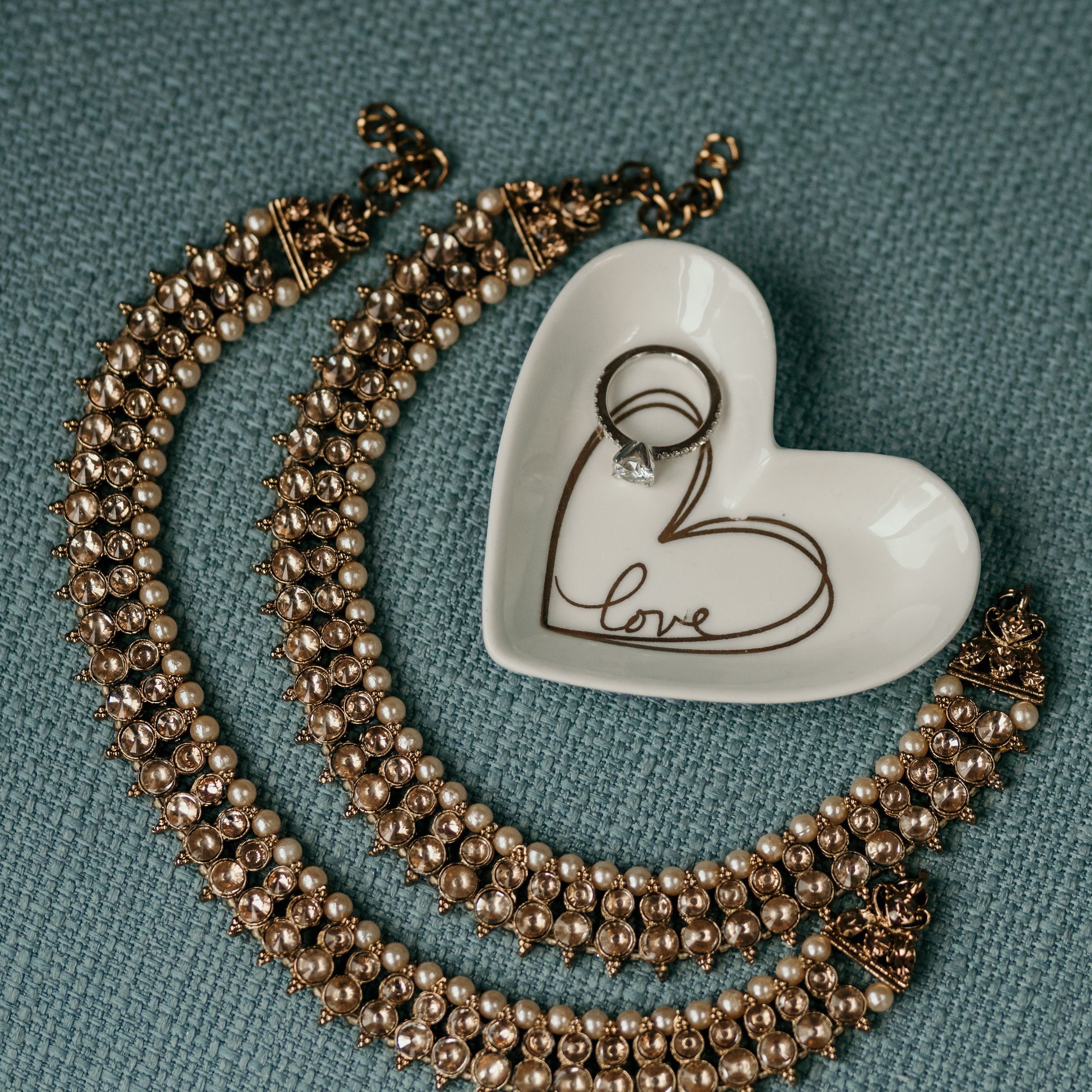 Gold beaded necklace and white heart-shaped trinket dish on gray fabric background.