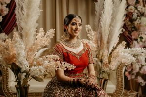 Bride in traditional red lehenga with gold jewelry seated among floral decorations.