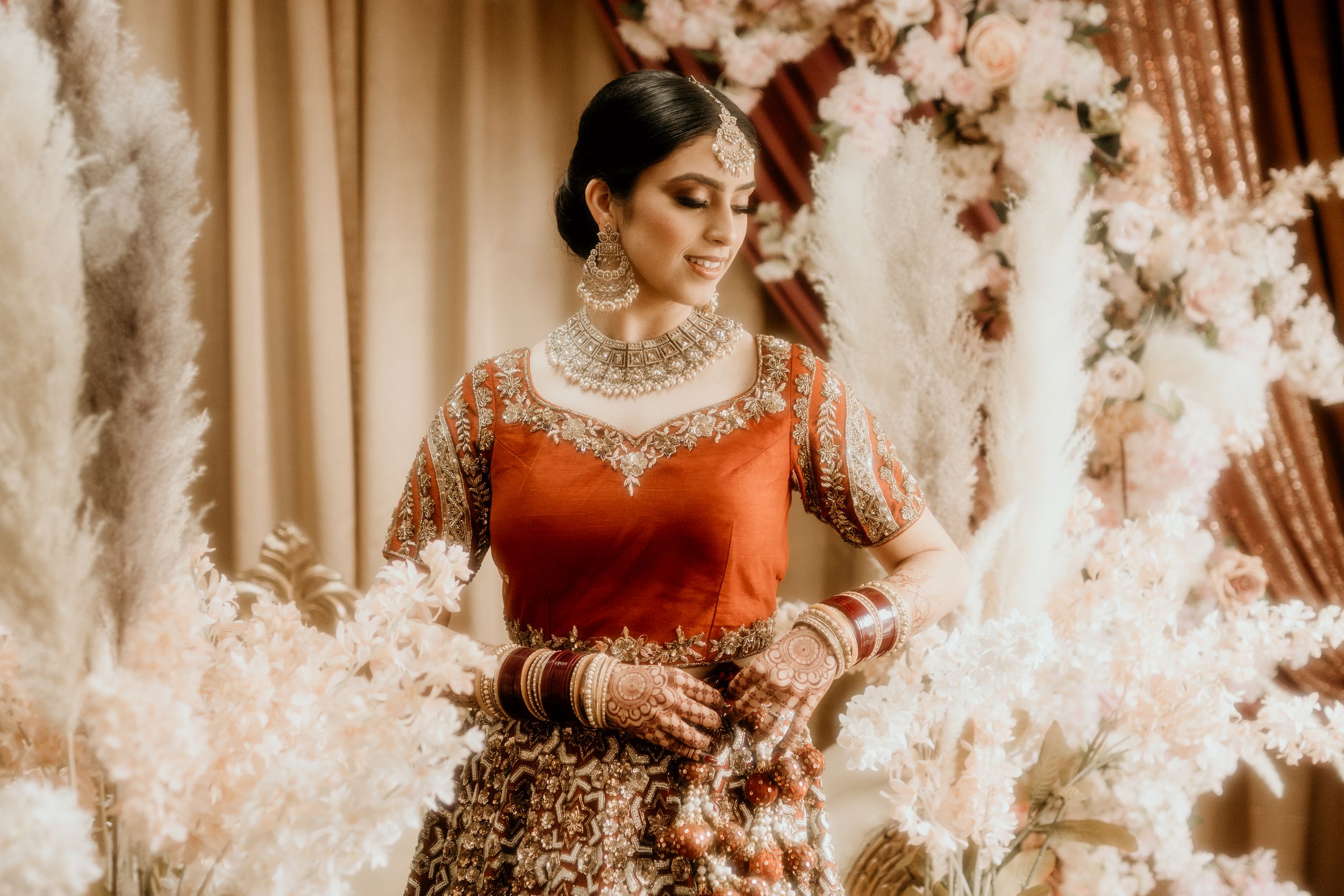 Indian bridal portrait in red and gold lehenga with white floral decor and warm lighting.