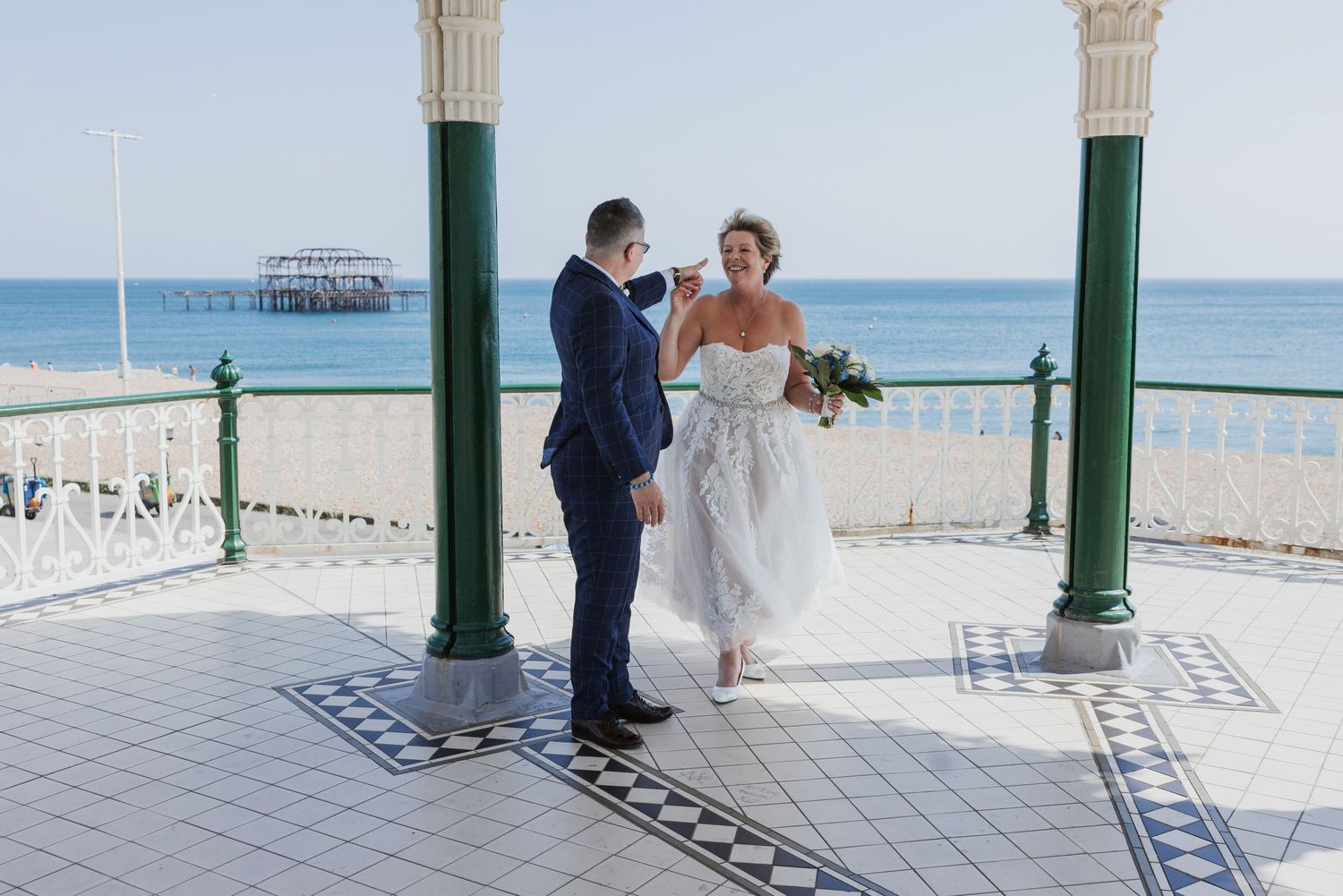 same sex couple dance in the Brighton Band stand on a very sunny day with the West Pier in the background