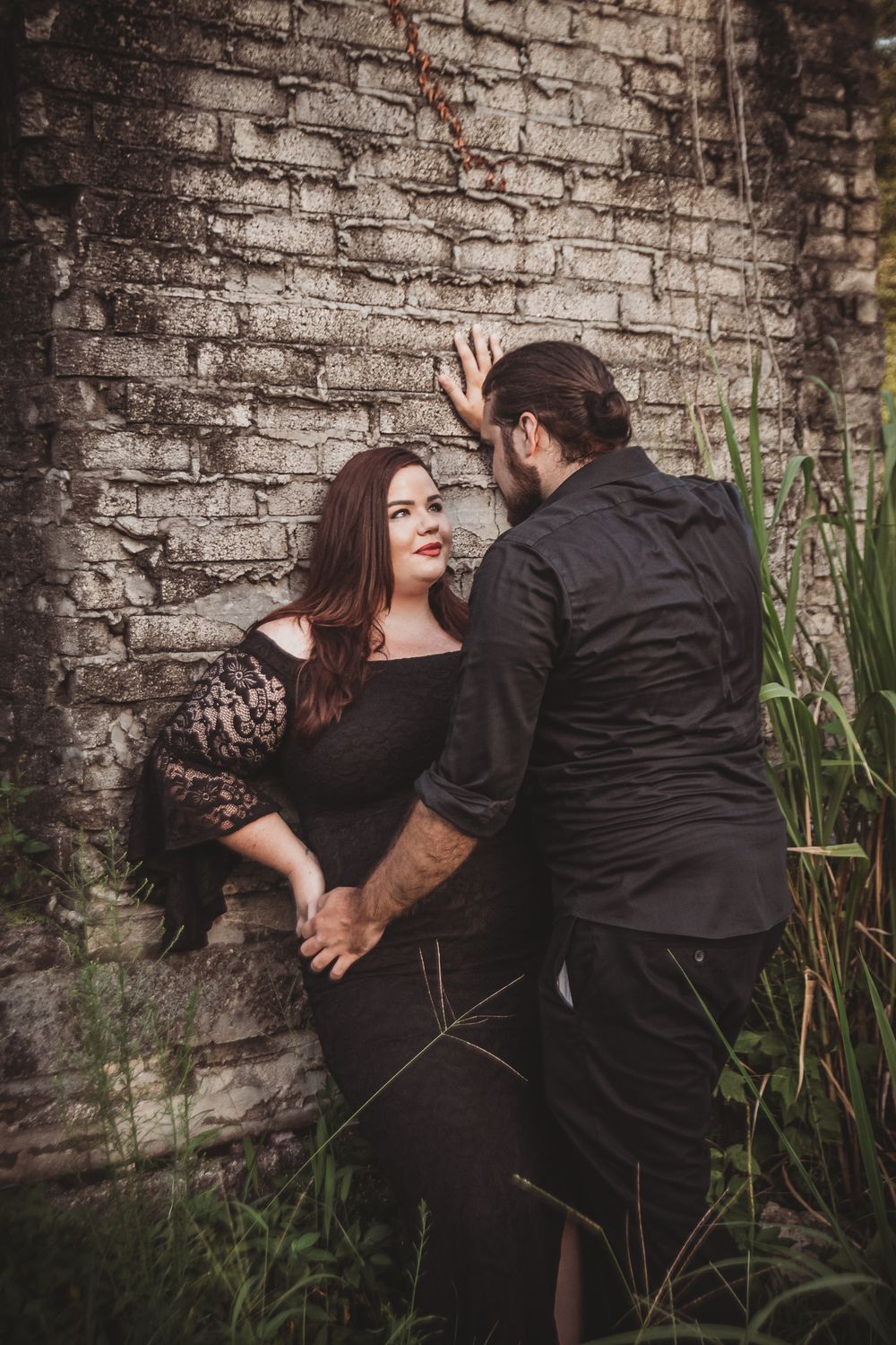 A romantic couple poses against a weathered brick wall surrounded by tall grass at dusk at the fireplace in riverbend park.