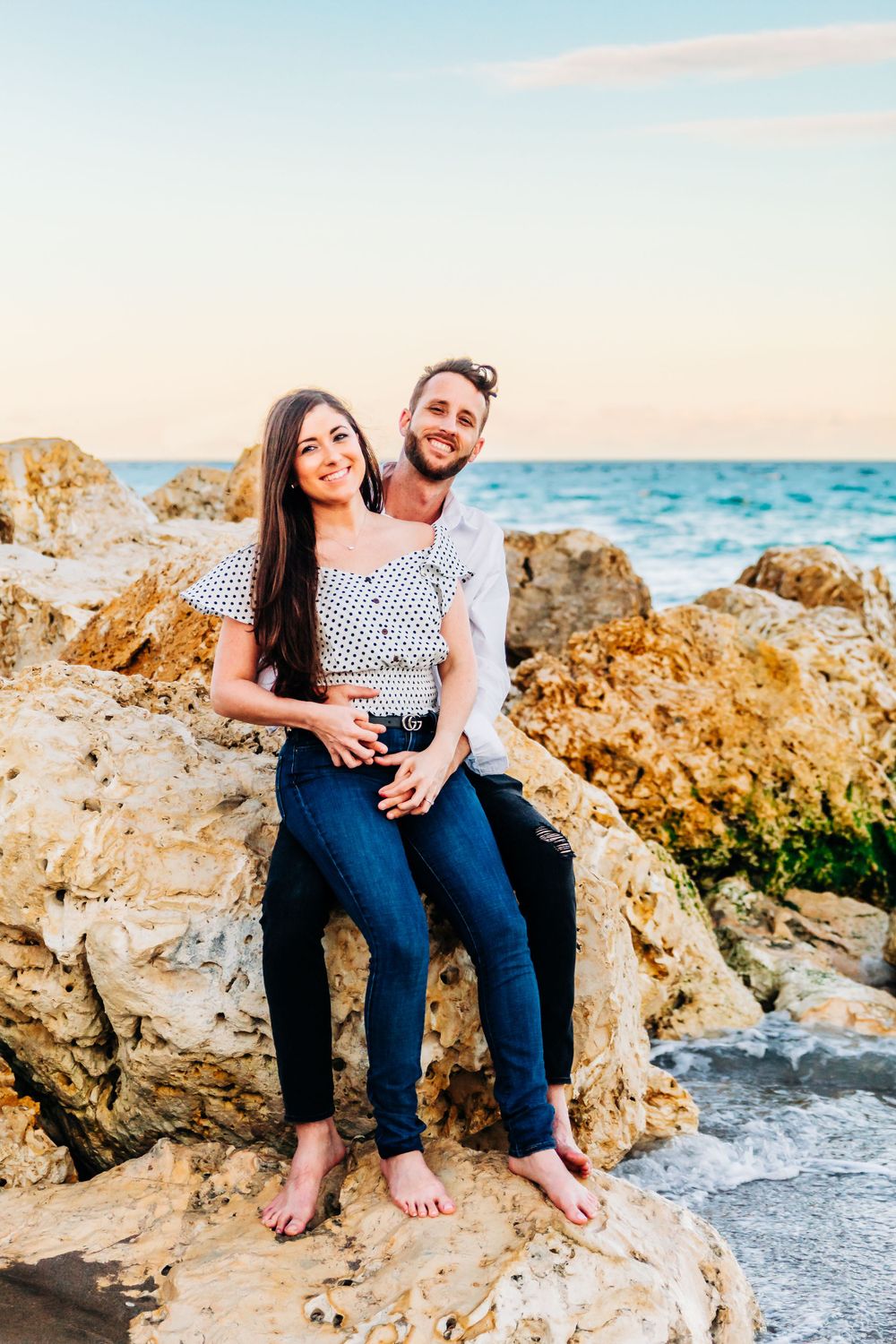 Happy couple sitting on coastal rocks with ocean view in coordinated casual outfits in palm beach Florida on the beach.