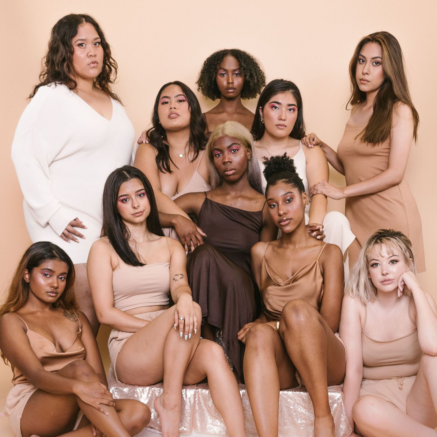 A group photo of 10 different women of colour, wearing a range of nude coloured garments and sitting together.