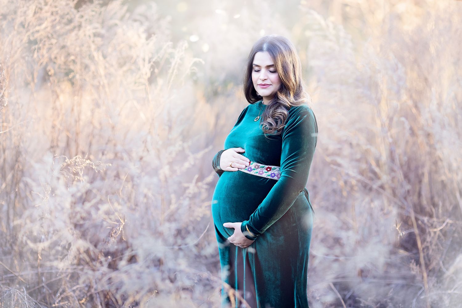 Pregnant woman in teal dress cradling baby bump in a golden field of tall dried grasses.