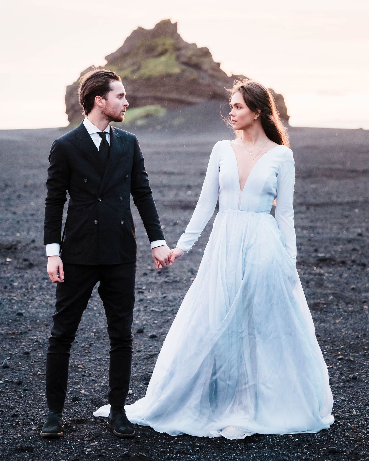 Couple in formal attire walking along a rocky volcanic landscape at sunset.