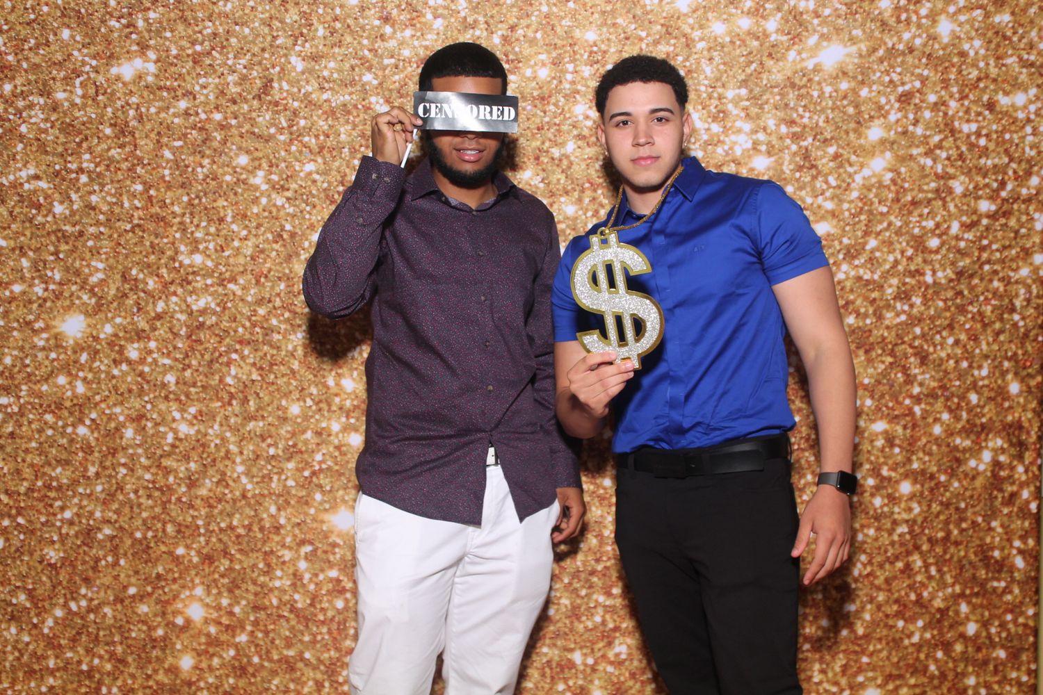 Two friends pose with fun props against a glittery gold background at a photo booth.
