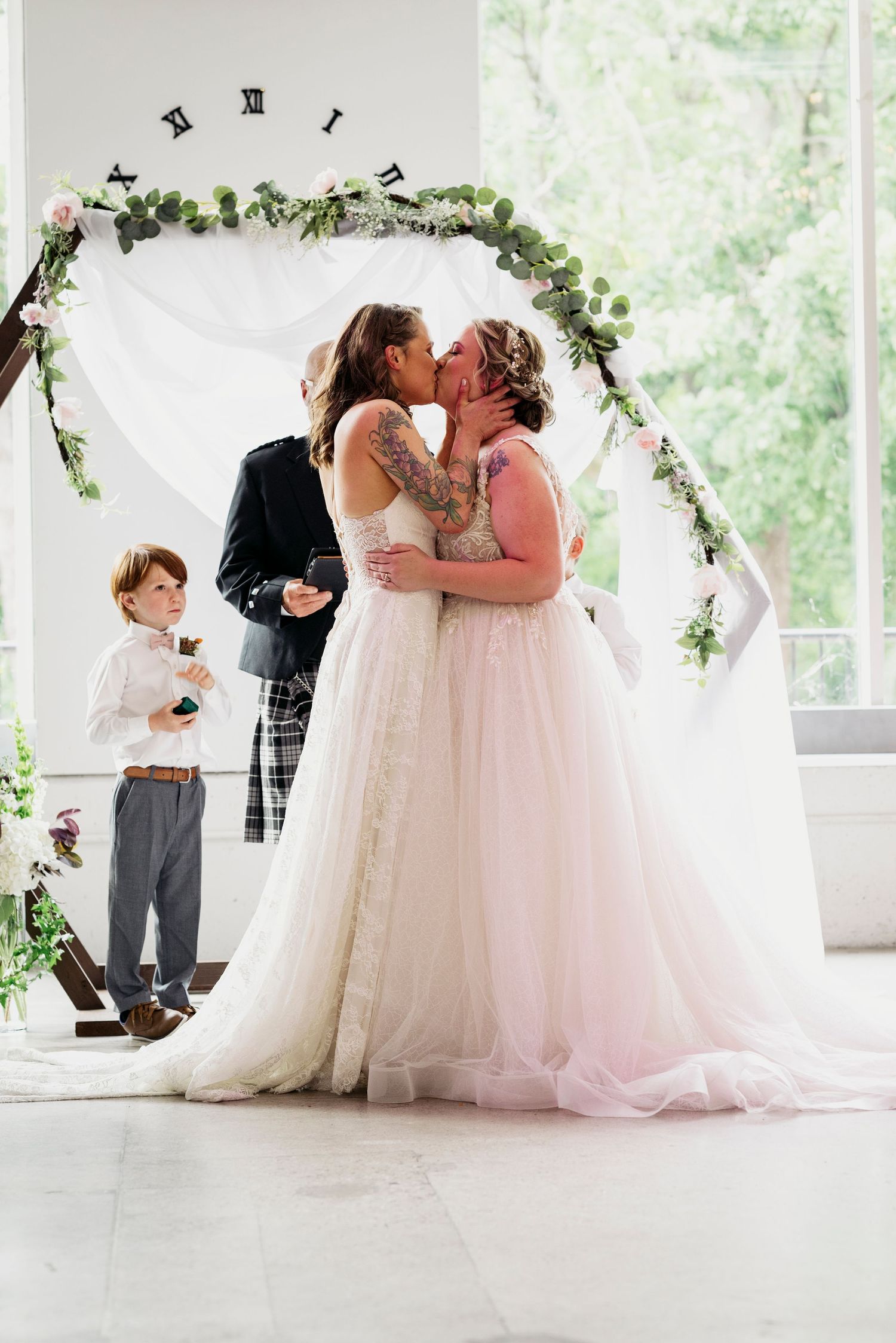 Two brides share a first kiss at their wedding ceremony beneath a floral arch.